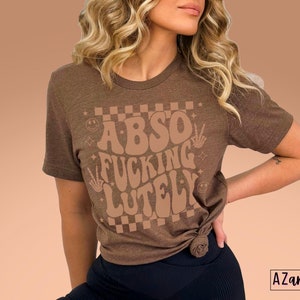 May include: A brown t-shirt with the text "Abso Fucking Lutely" in a retro font with a checkered background.