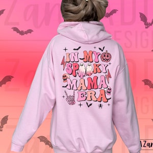 May include: A light pink hoodie with the text "IN MY SPOOKY MAMA ERA" in a retro font, along with Halloween-themed graphics like ghosts, a pumpkin, and a skull. The back of the hoodie features a design with bats and the brand name "Zand Addi Designs".