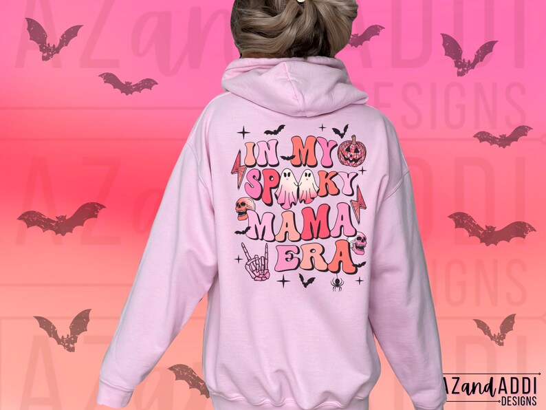May include: A light pink hoodie with the text "IN MY SPOOKY MAMA ERA" in a retro font, along with Halloween-themed graphics like ghosts, a pumpkin, and a skull. The back of the hoodie features a design with bats and the brand name "Zand Addi Designs".