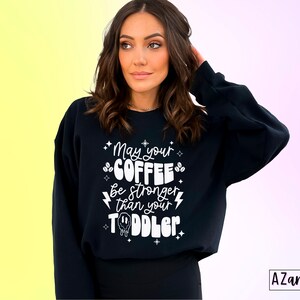 May include: Black sweatshirt with white text that reads "May your coffee be stronger than your toddler". The text is surrounded by small stars and coffee bean graphics.