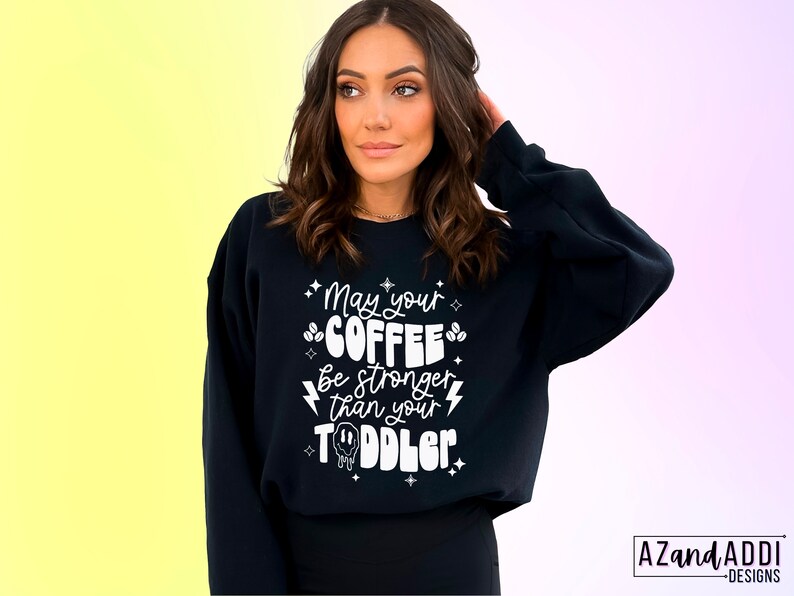 May include: Black sweatshirt with white text that reads "May your coffee be stronger than your toddler". The text is surrounded by small stars and coffee bean graphics.