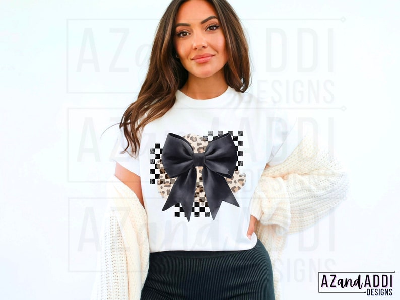 May include: White t-shirt with a black and white checkered bow with a leopard print design. The bow is centered on the shirt.