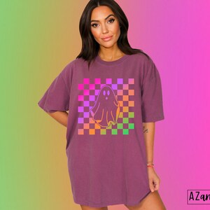 Op de afbeelding: Een bordeauxrood T-shirt met een spookafbeelding op een geruite achtergrond. De geest is wit omlijnd en het geruite patroon heeft roze, paarse, oranje en groene vierkanten. De tekst "AZandADDI DESIGNS" staat rechtsonder.