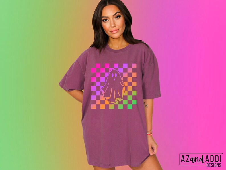 Op de afbeelding: Een bordeauxrood T-shirt met een spookafbeelding op een geruite achtergrond. De geest is wit omlijnd en het geruite patroon heeft roze, paarse, oranje en groene vierkanten. De tekst "AZandADDI DESIGNS" staat rechtsonder.