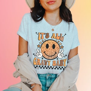 May include: A white t-shirt with a brown and orange smiley face with a leopard print design. The text "It's All Gravy Baby" is printed on the shirt.