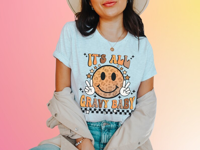 May include: A white t-shirt with a brown and orange smiley face with a leopard print design. The text "It's All Gravy Baby" is printed on the shirt.