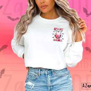 May include: White sweatshirt with a Halloween-themed graphic. The graphic features a pink and white ghost with the words "Spooky Mama" in pink and black. The background is a gradient of pink and orange with bat silhouettes.