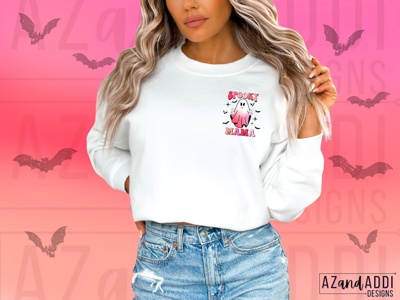 May include: White sweatshirt with a Halloween-themed graphic. The graphic features a pink and white ghost with the words "Spooky Mama" in pink and black. The background is a gradient of pink and orange with bat silhouettes.