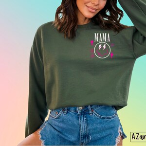 Pu&ograve; includere: Felpa verde con una grafica bianca di un viso sorridente con fulmini e la parola "MAMA" sopra.