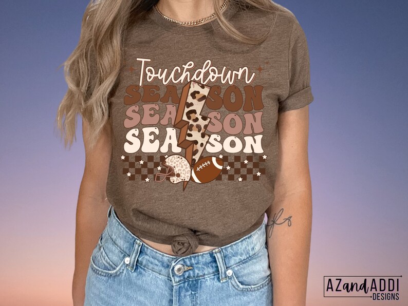 May include: A brown t-shirt with the text "Touchdown Season" in a retro font. The text is layered with a leopard print and a lightning bolt graphic. The shirt also features a football and a football helmet.