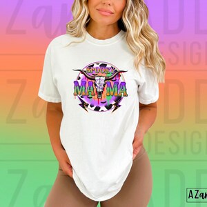 May include: White t-shirt with a graphic design featuring a skull, horns, and the words "Spooky Mama" in purple and green. The design includes a cow print and lightning bolts. The shirt is a casual style.