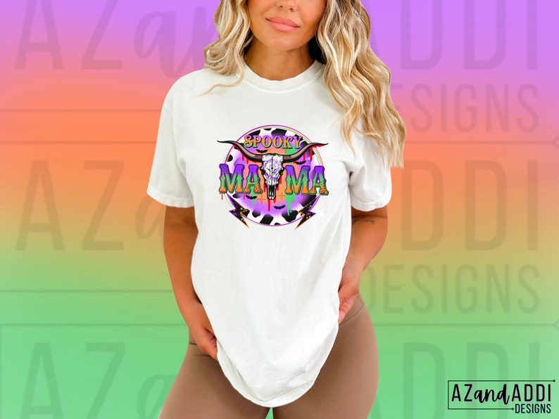 May include: White t-shirt with a graphic design featuring a skull, horns, and the words "Spooky Mama" in purple and green. The design includes a cow print and lightning bolts. The shirt is a casual style.