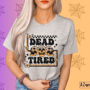 Op de afbeelding: Een grijs T-shirt met een grafische print van drie schedels met luipaardprint en de tekst "Dead Tired".