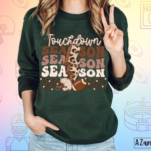 May include: Green sweatshirt with the text "Touchdown Season" in brown, pink, and white lettering. The text is layered with a brown and black leopard print and a lightning bolt graphic.