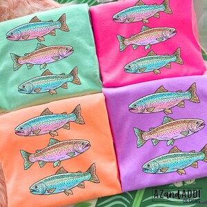 Puede incluir: Varias camisetas de colores brillantes en tonos rosa, verde, naranja y morado, cada una con una ilustraci&oacute;n detallada de una trucha. Los peces est&aacute;n representados con escamas azules, rosas y marrones. La camiseta inferior derecha tiene el texto "AZandADDI DESIGNS".