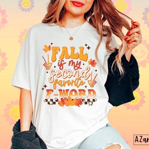 Pu&ograve; includere: T-shirt color crema con la scritta "FALL is my second favorite F WORD" in arancione, marrone e bianco. Il design include foglie autunnali, una zucca e una mano scheletrica. Fa parte di una collezione a tema autunnale.