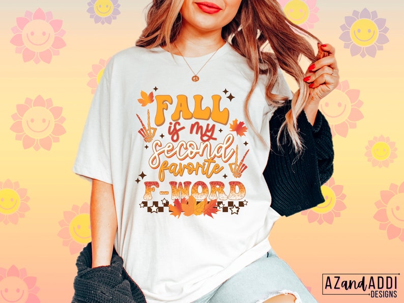 Pu&ograve; includere: T-shirt color crema con la scritta "FALL is my second favorite F WORD" in arancione, marrone e bianco. Il design include foglie autunnali, una zucca e una mano scheletrica. Fa parte di una collezione a tema autunnale.
