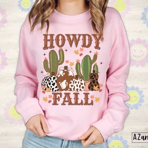 May include: A light pink sweatshirt with a graphic design featuring the words "HOWDY FALL" in brown lettering. The design includes cacti, pumpkins with cow print and fall leaves. The sweatshirt is a long-sleeved crew neck.