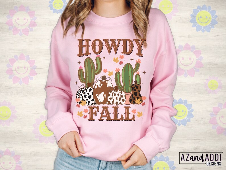 May include: A light pink sweatshirt with a graphic design featuring the words "HOWDY FALL" in brown lettering. The design includes cacti, pumpkins with cow print and fall leaves. The sweatshirt is a long-sleeved crew neck.