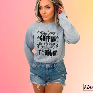 May include: A gray sweatshirt with the text "May your coffee be stronger than your toddler" in black lettering. The text is surrounded by small stars and lightning bolts.