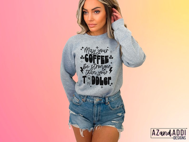 May include: A gray sweatshirt with the text "May your coffee be stronger than your toddler" in black lettering. The text is surrounded by small stars and lightning bolts.
