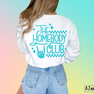 Puede incluir: Camiseta blanca de manga larga con un gr&aacute;fico azul turquesa que dice "The Homebody Club" con una mano de esqueleto haciendo un signo de rock and roll. El gr&aacute;fico est&aacute; rodeado de peque&ntilde;as estrellas y un patr&oacute;n a cuadros.