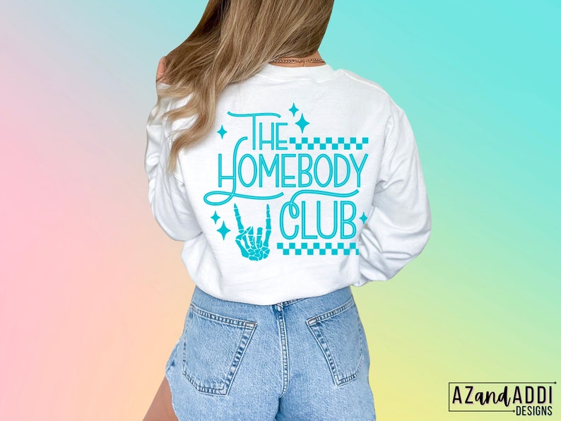 Puede incluir: Camiseta blanca de manga larga con un gr&aacute;fico azul turquesa que dice "The Homebody Club" con una mano de esqueleto haciendo un signo de rock and roll. El gr&aacute;fico est&aacute; rodeado de peque&ntilde;as estrellas y un patr&oacute;n a cuadros.