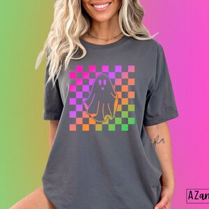 Op de afbeelding: Donkergrijs T-shirt met een kleurrijk ruitpatroon en een spookafbeelding. Het spook is omlijnd in oranje en het ruitpatroon bevat roze, paarse, oranje en groene vierkanten. De tekst "AZandAddi DESIGNS" staat rechtsonder.