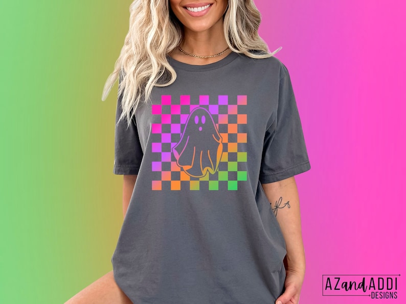 Op de afbeelding: Donkergrijs T-shirt met een kleurrijk ruitpatroon en een spookafbeelding. Het spook is omlijnd in oranje en het ruitpatroon bevat roze, paarse, oranje en groene vierkanten. De tekst "AZandAddi DESIGNS" staat rechtsonder.