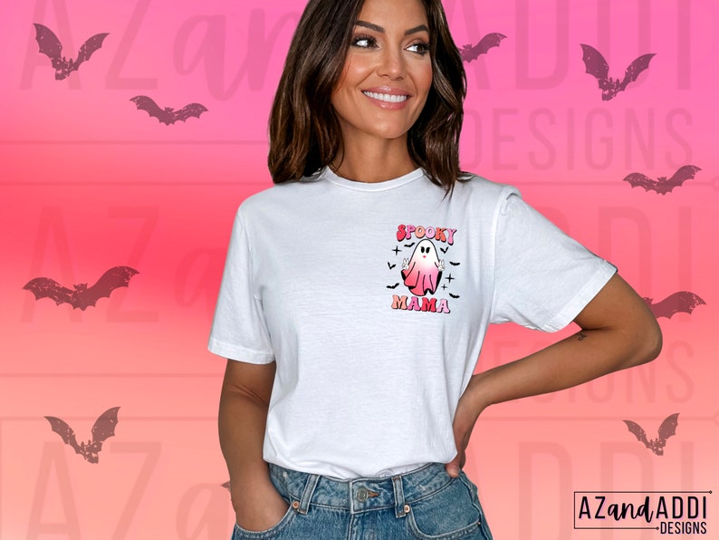 May include: White t-shirt with a "Spooky Mama" graphic featuring a ghost in pink and white, with bats and crosses. The design is in a retro style. The shirt is a short-sleeved crew neck.