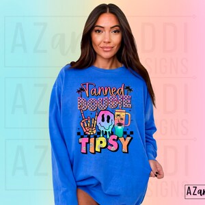 May include: A royal blue sweatshirt with a colourful graphic design that says "Tanned Bougie Tipsy" with a smiley face, palm trees, and a chequered pattern.