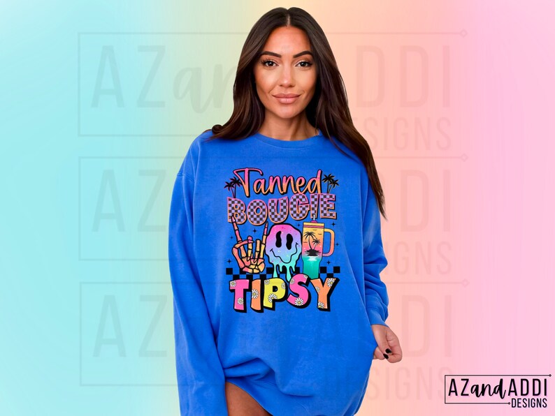 May include: A royal blue sweatshirt with a colourful graphic design that says "Tanned Bougie Tipsy" with a smiley face, palm trees, and a chequered pattern.