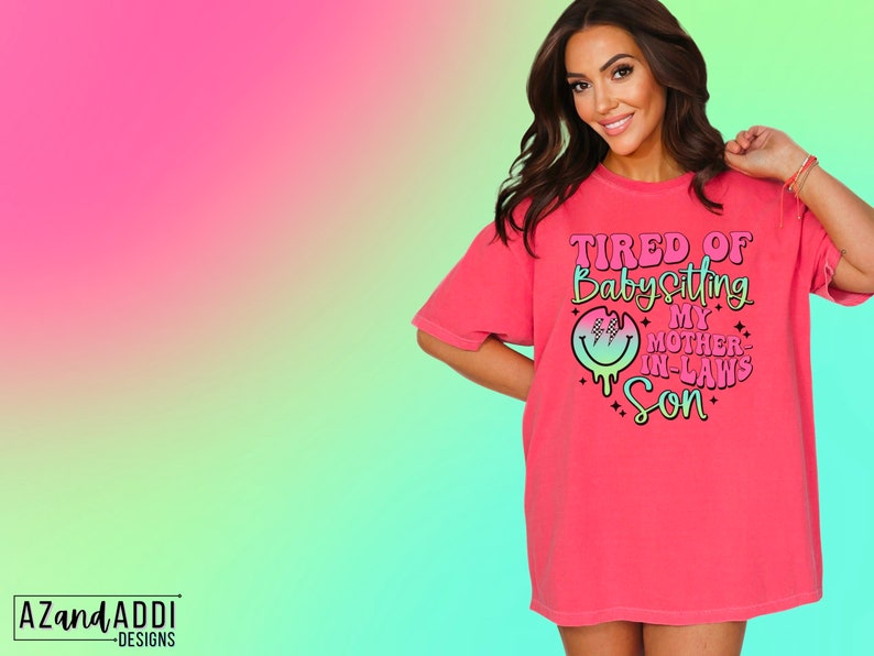 Puede incluir: Una camiseta rosa con el texto "TIRED OF BABYSITTING MY MOTHER-IN-LAWS SON" en letras rosa y negras. Una cara sonriente con un degradado arco&iacute;ris y un patr&oacute;n de cuadros negros est&aacute; por encima del texto.