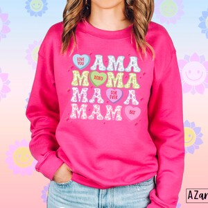 May include: Pink sweatshirt with a graphic design of the word "AMA" repeated four times in different colors and fonts. Each "AMA" is surrounded by a heart with a different message on each heart: "LOVE YOU", "XOXO", "FOR EVER", and "BFF".