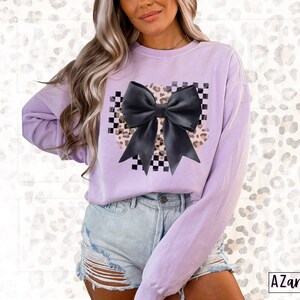 May include: A lavender sweatshirt with a black bow on a checkered and leopard print background.
