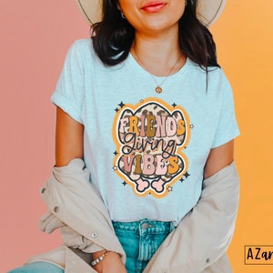 May include: Light gray t-shirt with rolled sleeves featuring the text "FRIENDS Giving VIBES" in a retro design. The graphic includes a leopard print and star accents. The shirt is part of a clothing collection.