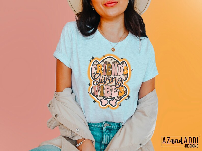 May include: Light gray t-shirt with rolled sleeves featuring the text "FRIENDS Giving VIBES" in a retro design. The graphic includes a leopard print and star accents. The shirt is part of a clothing collection.