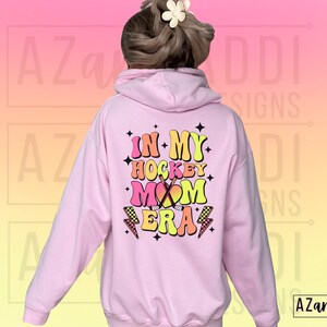 May include: Pink hoodie with a graphic that says "In My Hockey Mom Era" in colorful retro lettering with a hockey stick and lightning bolt accents.