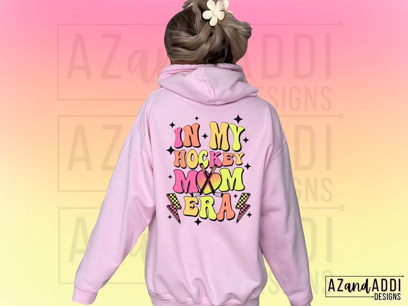 May include: Pink hoodie with a graphic that says "In My Hockey Mom Era" in colorful retro lettering with a hockey stick and lightning bolt accents.