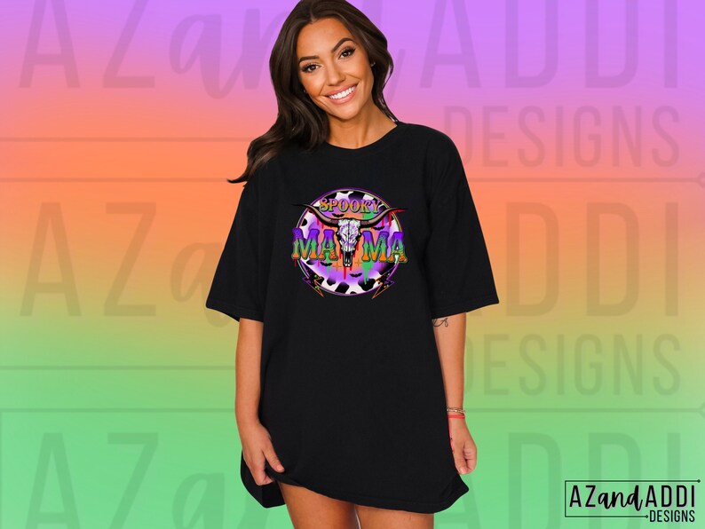 May include: Black t-shirt featuring the text "Spooky Mama" in a colourful graphic with a skull and horns design. The graphic has purple, green, and orange accents. The t-shirt has a relaxed fit.