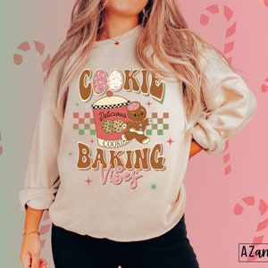 May include: A beige sweatshirt with a retro design that reads "Cookie Baking Vibes." The graphic includes a cookie jar, gingerbread man, and decorated eggs. The design is in brown, pink, and green colors. The sweatshirt is a cozy, casual style.