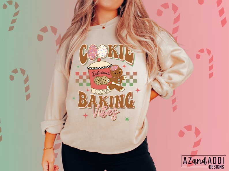 May include: A beige sweatshirt with a retro design that reads "Cookie Baking Vibes." The graphic includes a cookie jar, gingerbread man, and decorated eggs. The design is in brown, pink, and green colors. The sweatshirt is a cozy, casual style.