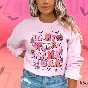 May include: A light pink sweatshirt with the text "IN MY SPOOKY MAMA ERA" in a retro font. The design includes bats, ghosts, a pumpkin, a skull, and a skeleton hand. The sweatshirt has long sleeves and a crew neck.