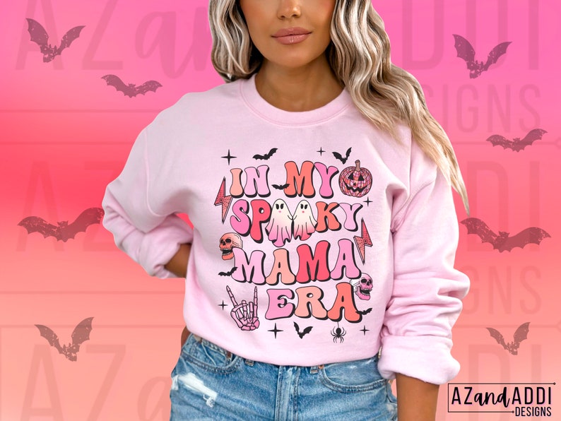 May include: A light pink sweatshirt with the text "IN MY SPOOKY MAMA ERA" in a retro font. The design includes bats, ghosts, a pumpkin, a skull, and a skeleton hand. The sweatshirt has long sleeves and a crew neck.
