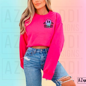 May include: A pink cropped sweatshirt featuring a graphic of a melting smiley face with palm trees and a checkered pattern. The sweatshirt is being worn by a person with long blonde hair.