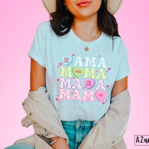 May include: A light blue t-shirt with a graphic design of the word "MAMA" spelled out with candy hearts. The hearts have different colors and sayings like "Love You", "XOXO", "For Ever", and "Bet".