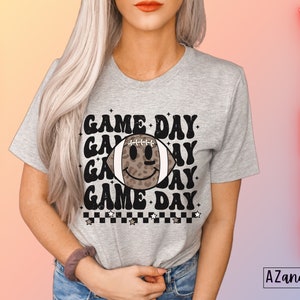 May include: A grey t-shirt featuring the text "GAME DAY" repeated, with a football graphic in the middle. The football has a leopard print design and a smiley face. The words "FOOTBALL" and "VIBES" are written vertically on either side of the shirt.