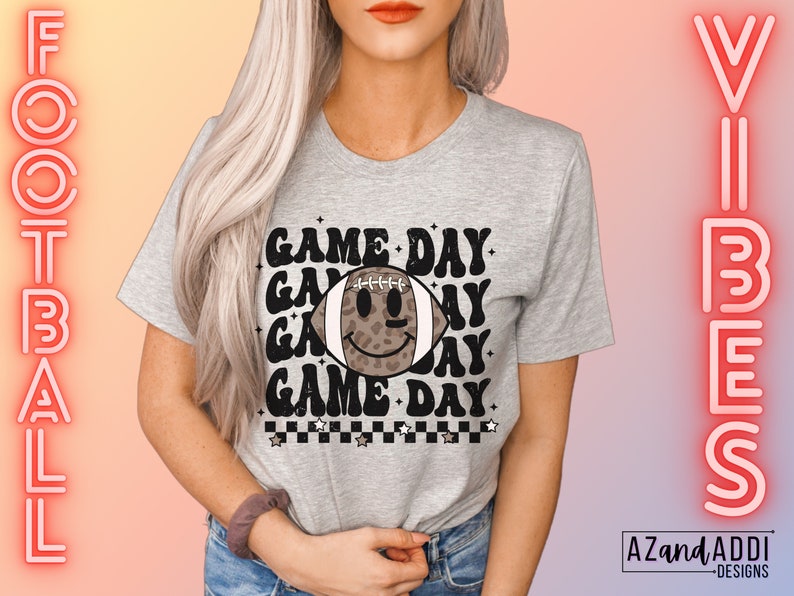 May include: A grey t-shirt featuring the text "GAME DAY" repeated, with a football graphic in the middle. The football has a leopard print design and a smiley face. The words "FOOTBALL" and "VIBES" are written vertically on either side of the shirt.