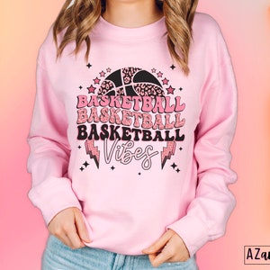 May include: A light pink sweatshirt with a basketball-themed graphic. The graphic includes the word "Basketball" repeated three times, a basketball, stars, and lightning bolts. The word "Vibes" is also included.