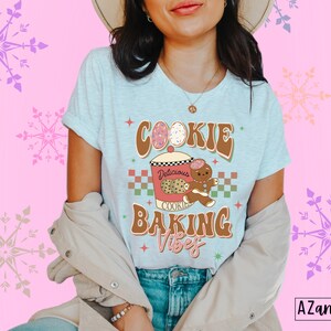 May include: Light blue t-shirt with the words "COOKIE BAKING VIBES" in brown and green retro font. The graphic includes a cookie jar, gingerbread man, and cookies. The shirt is worn by a person.